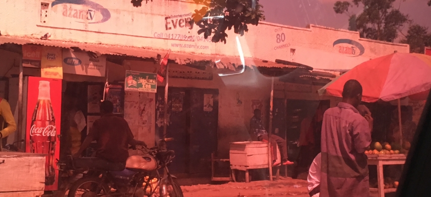 photo taken from inside a car looking out at a road side shop. on the left is a big sign with a coke bottle on it and the writing coca cola. one can see a motorbike on the left and a person walking by on the right. The picture has red tint to it due to the coloring of the car windows. Behind the shop are tropical plants