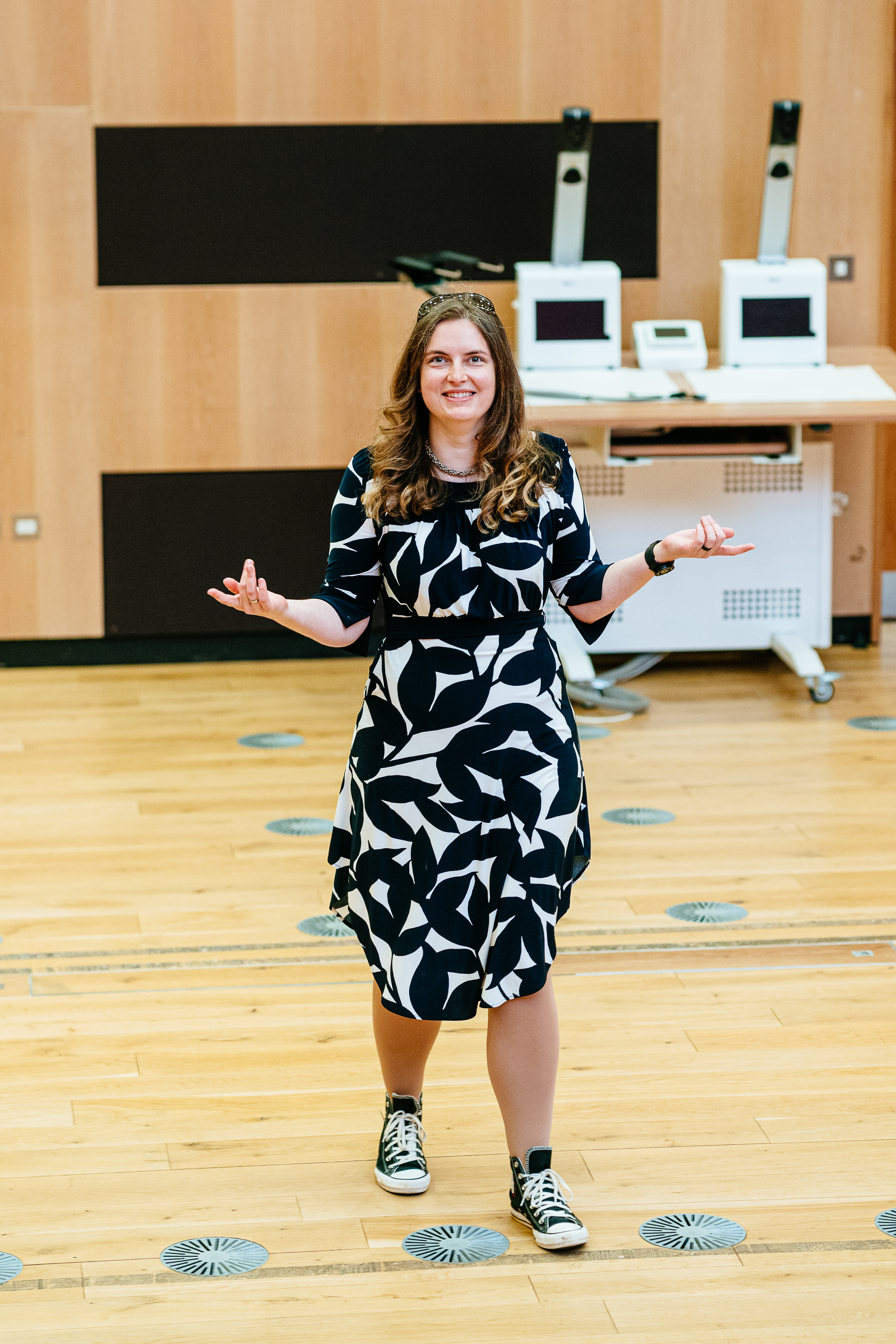 Anica is standing large lecture hall. she wears a black and white dress with black and white Converse shoes. She holds her arms out to the side and is mid step smiling