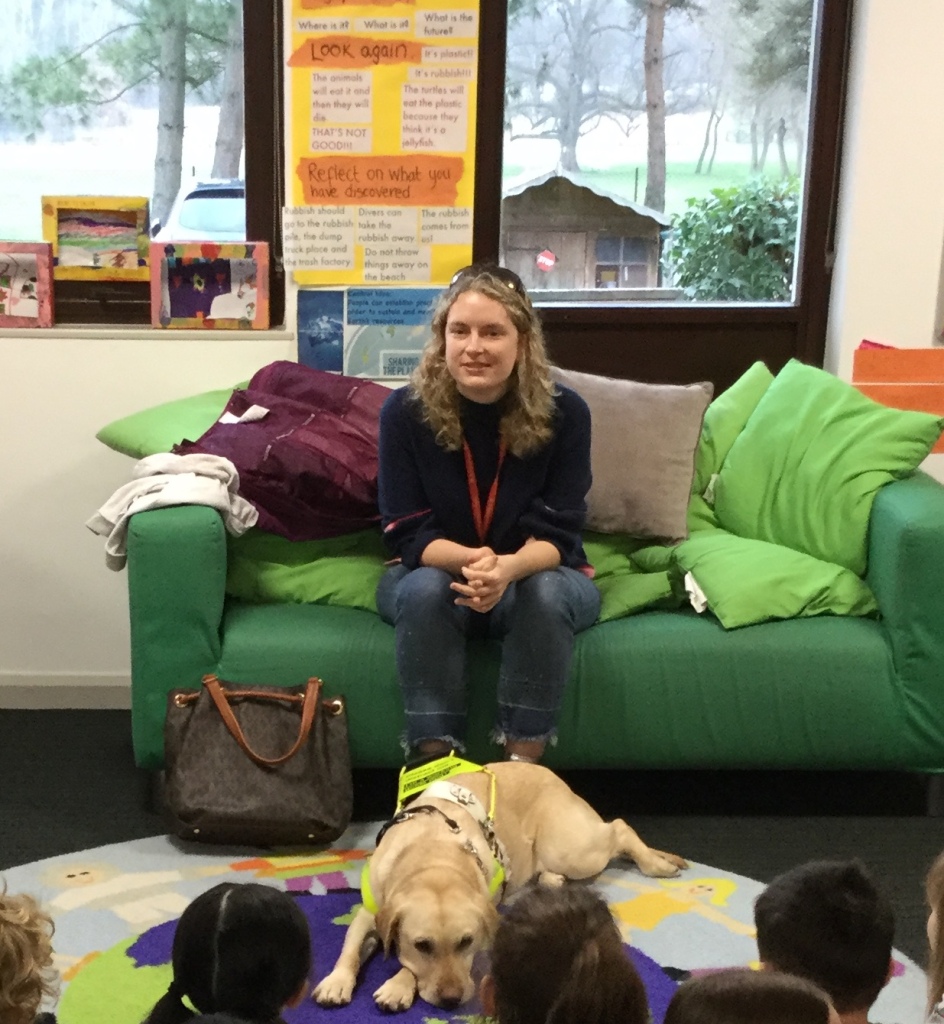 Anica sits on a sofa in a primary school classroom. Lassie her guide dog lies on the floor in front of her