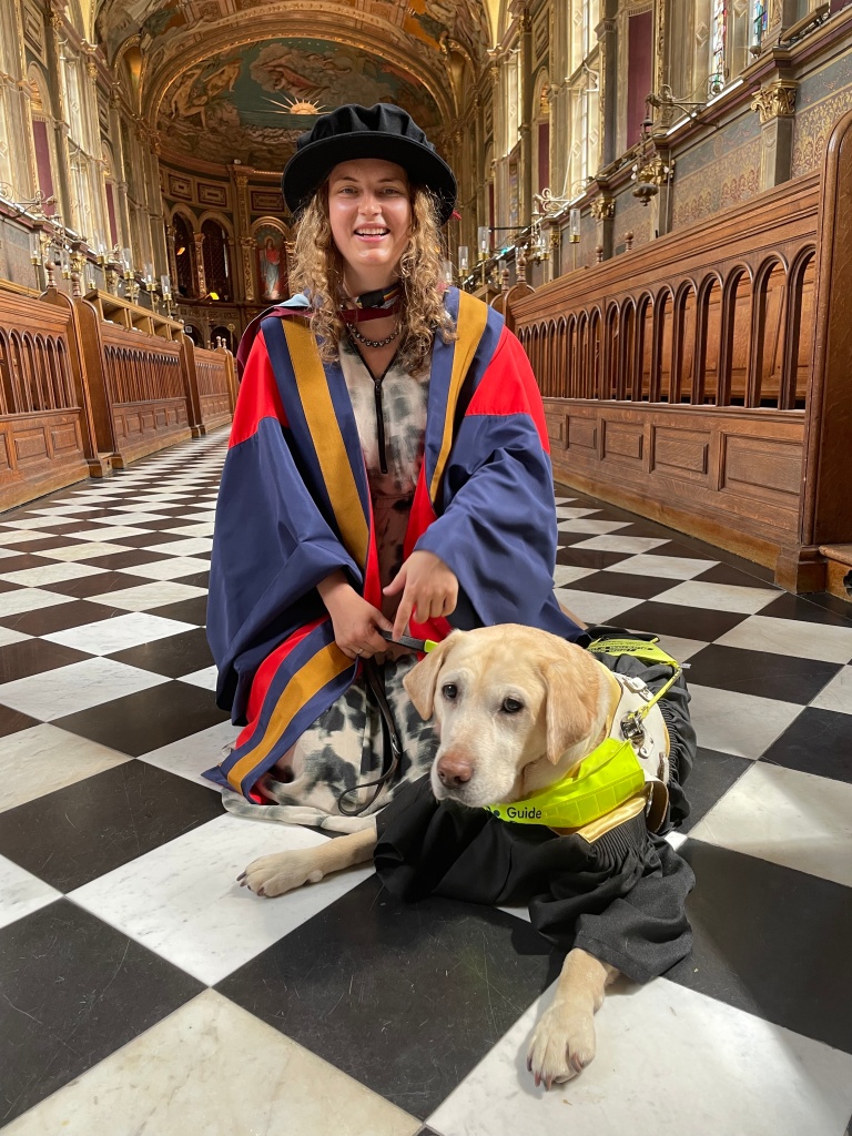 Anica Zeyen, a white woman with dark blonde curly hair is kneeling on the floor wearing a blue, yellow and red academic gown and doctoral round hat. Next to her, is her former guide dog Lassie lying down wearing a black and gold academic gown underneath her guide dog harness. They are in the Chapel of Royal Holloway, a beautifully decorated Victorian chapel. 
