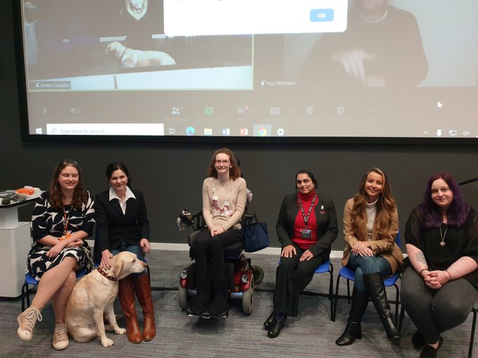 photo of a panel with six women. From left to right: Anica Zeyen and her guide dog Lassie, Oana Branzei, Hannah Deakin, Avril Coelho, Kelly Given and Lucy Matthews. The background shows a screen with the broadcast of the panel